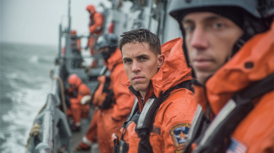 Coast Guard members in orange rain gear on a patrol boat during stormy weather, with one man looking directly at the camera