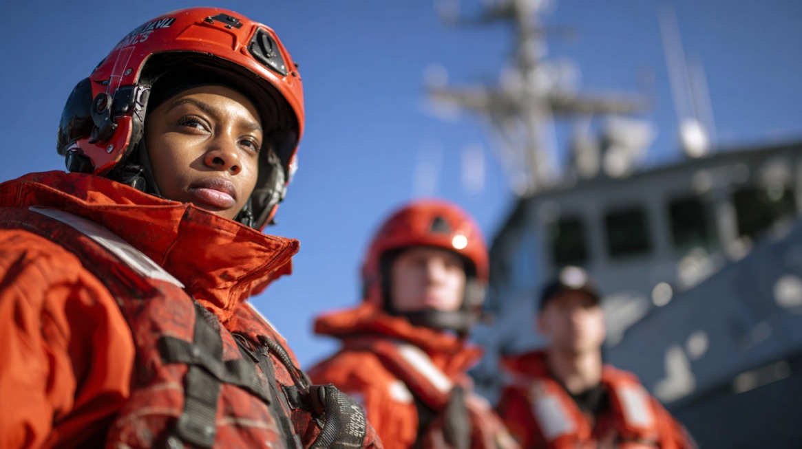 Coast Guard reservist in orange safety gear with helmet, standing in front of a ship with fellow team members in the background