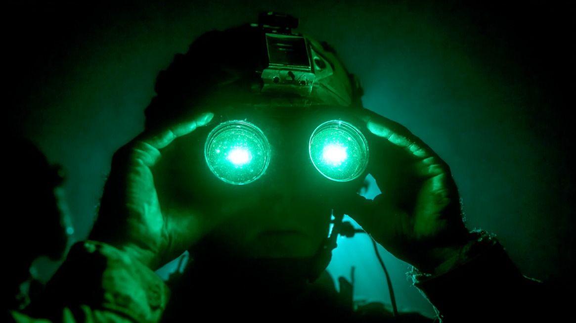 Soldier using night vision goggles in a dark environment, with lenses glowing green