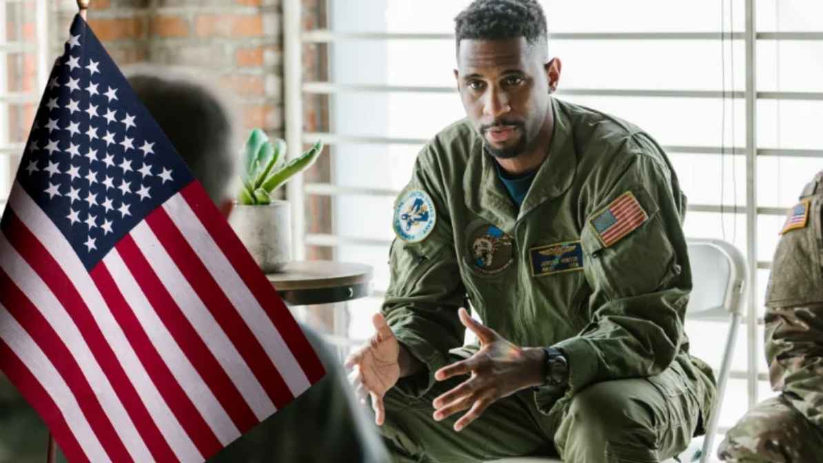 A U.S. military servicemember in uniform sits and speaks during a discussion, with an American flag in the foreground