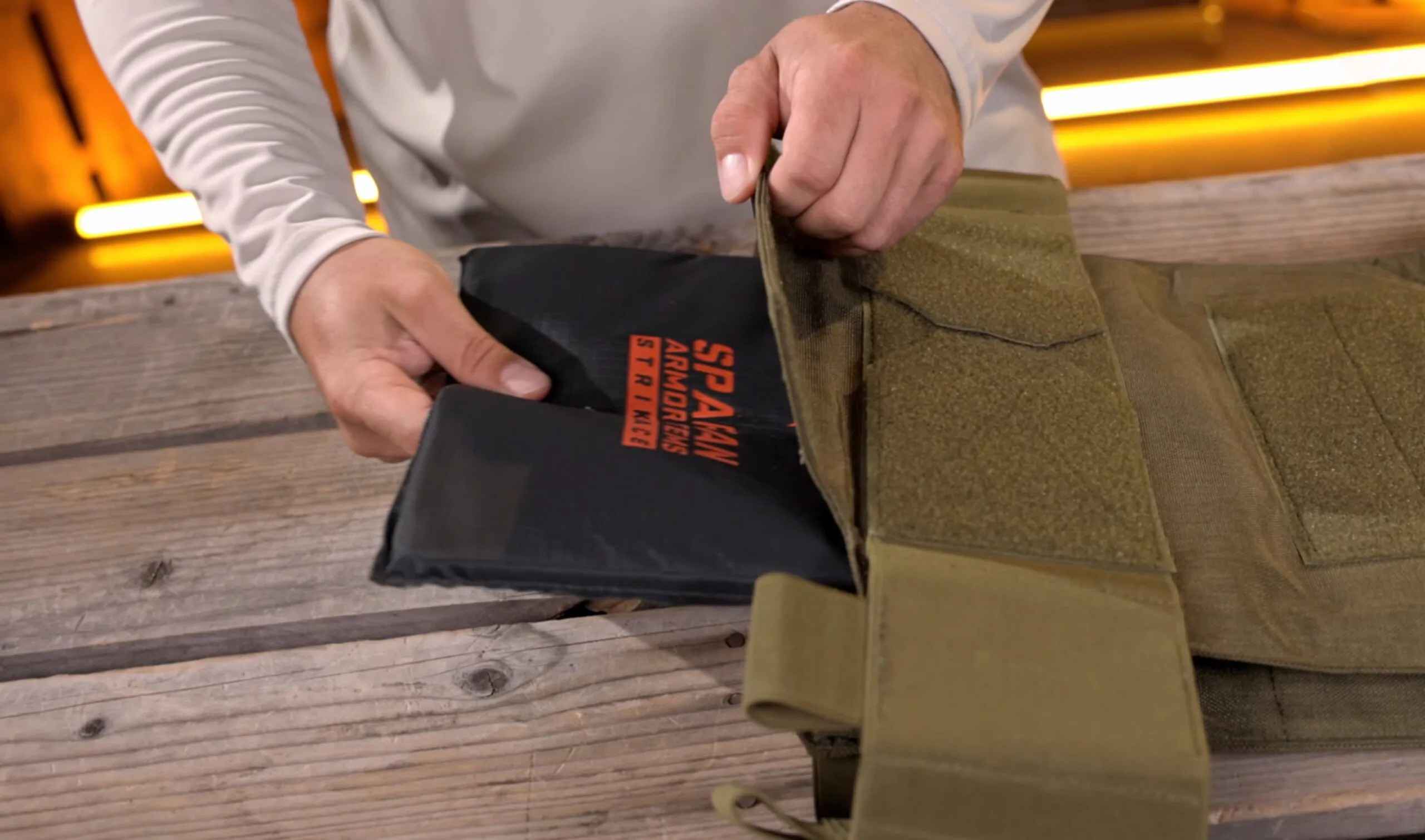 Hands inserting a Spartan Armor Systems ballistic plate into a tan plate carrier, displayed on a wooden surface