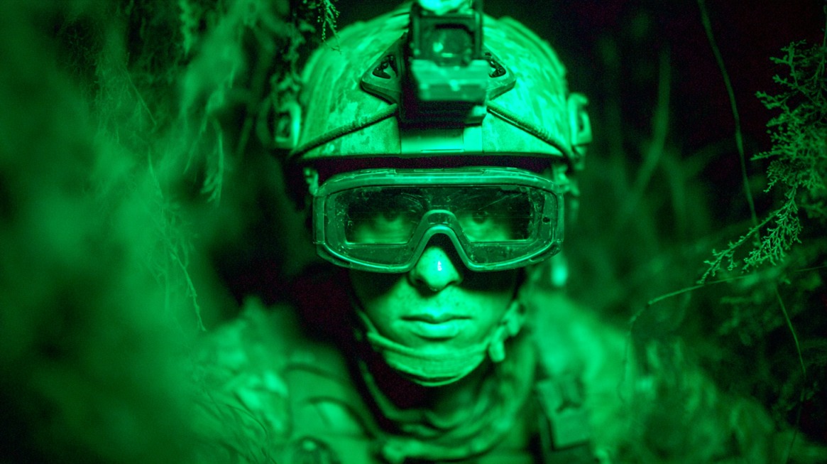 Soldier wearing early-model night vision gear in a dark, forested area, illuminated in green