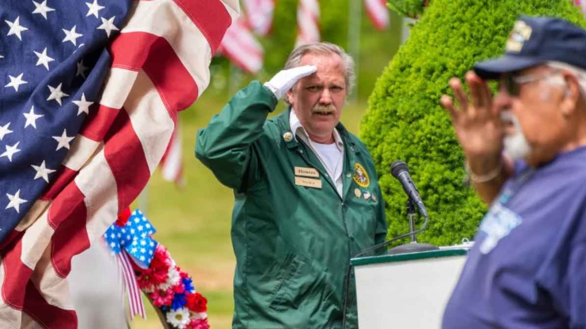 Two veterans salute during a ceremony with American flags in the background and one standing at a podium
