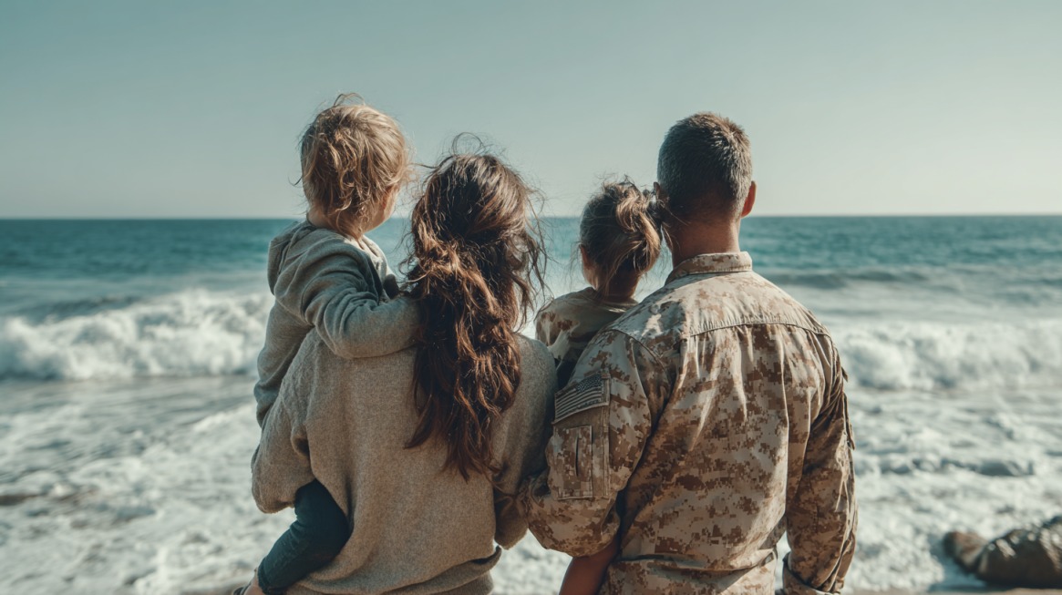 Military family of four facing the ocean, with the service member in uniform and two children held by their parents