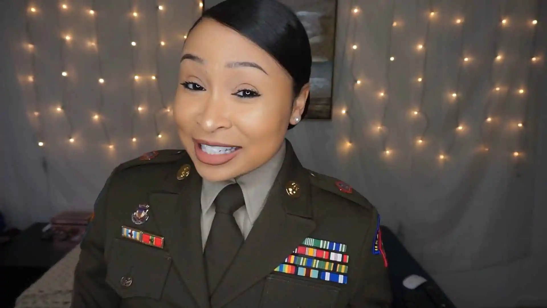 A U.S. Army soldier in the Army Green Service Uniform (AGSU), smiling confidently with decorative lights in the background