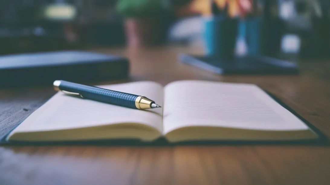 Close-up of an open notebook with a pen resting on its pages, on a wooden desk with blurred background items