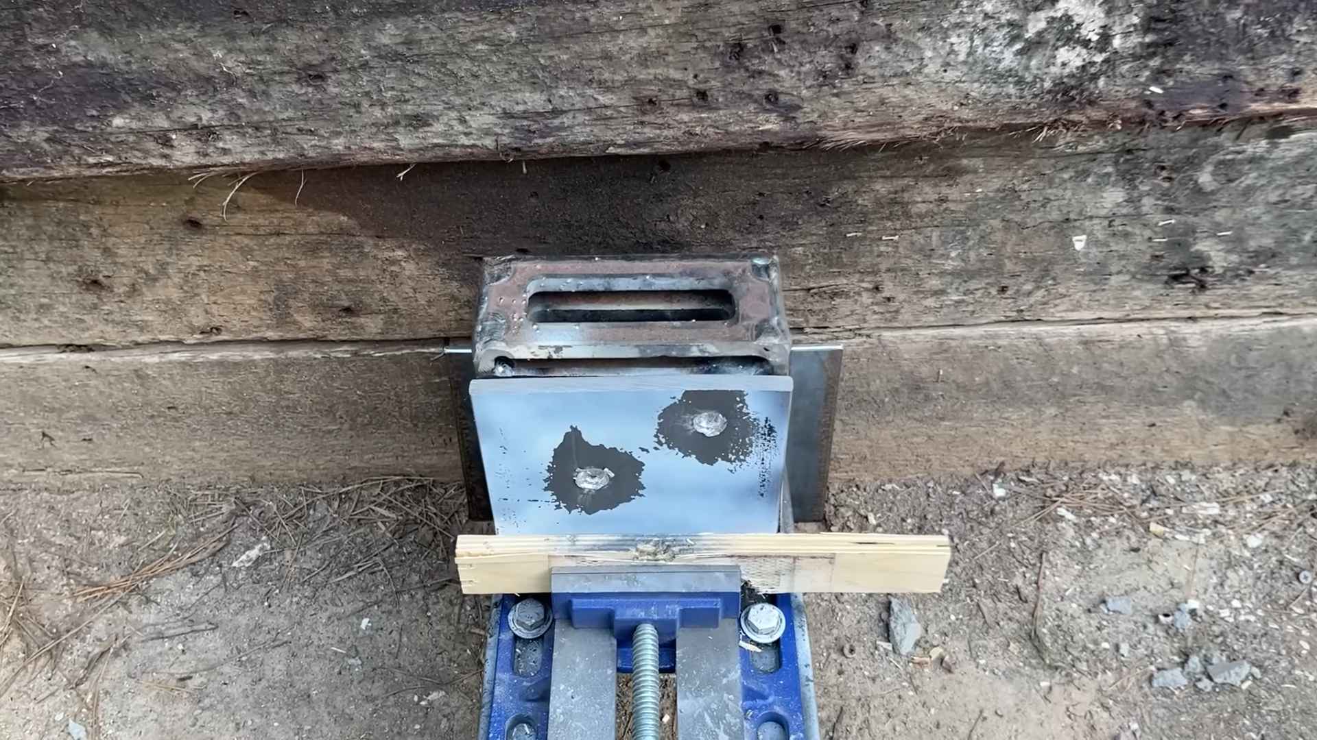 Close-up of a steel plate with bullet impact marks from ballistic testing, mounted against a wooden backstop