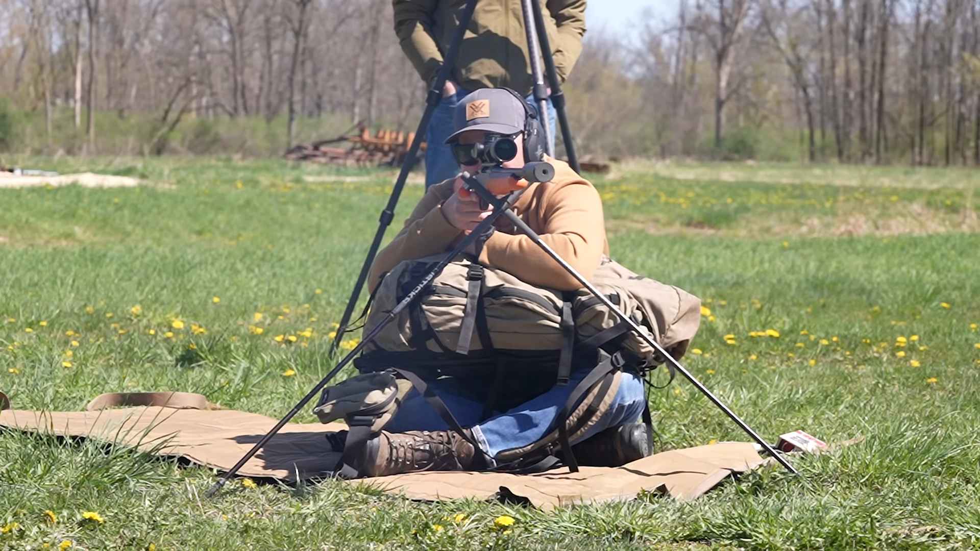 Shooter in outdoor setting using a rifle with a bipod and scope, aiming from a supported seated position on a shooting mat