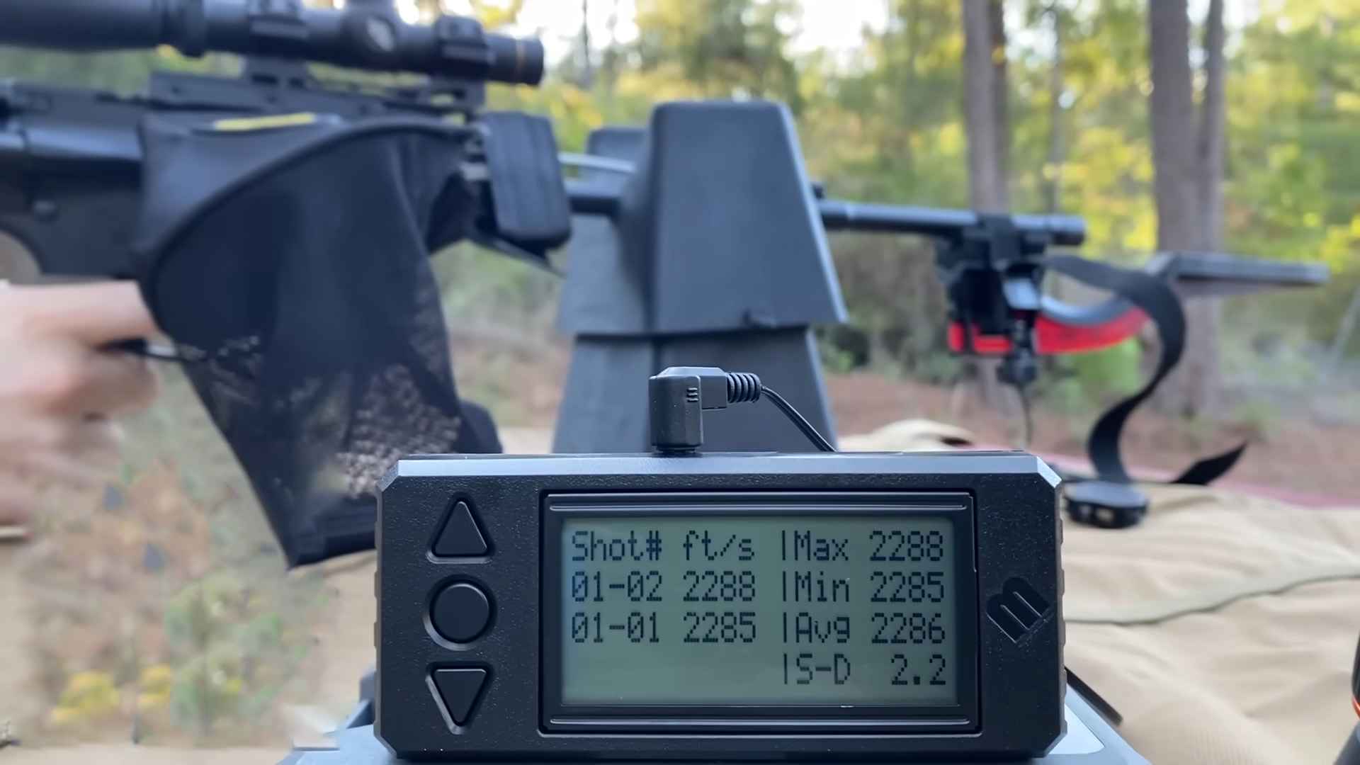 Close-up of a chronograph displaying bullet velocity data in front of a rifle setup at a shooting range