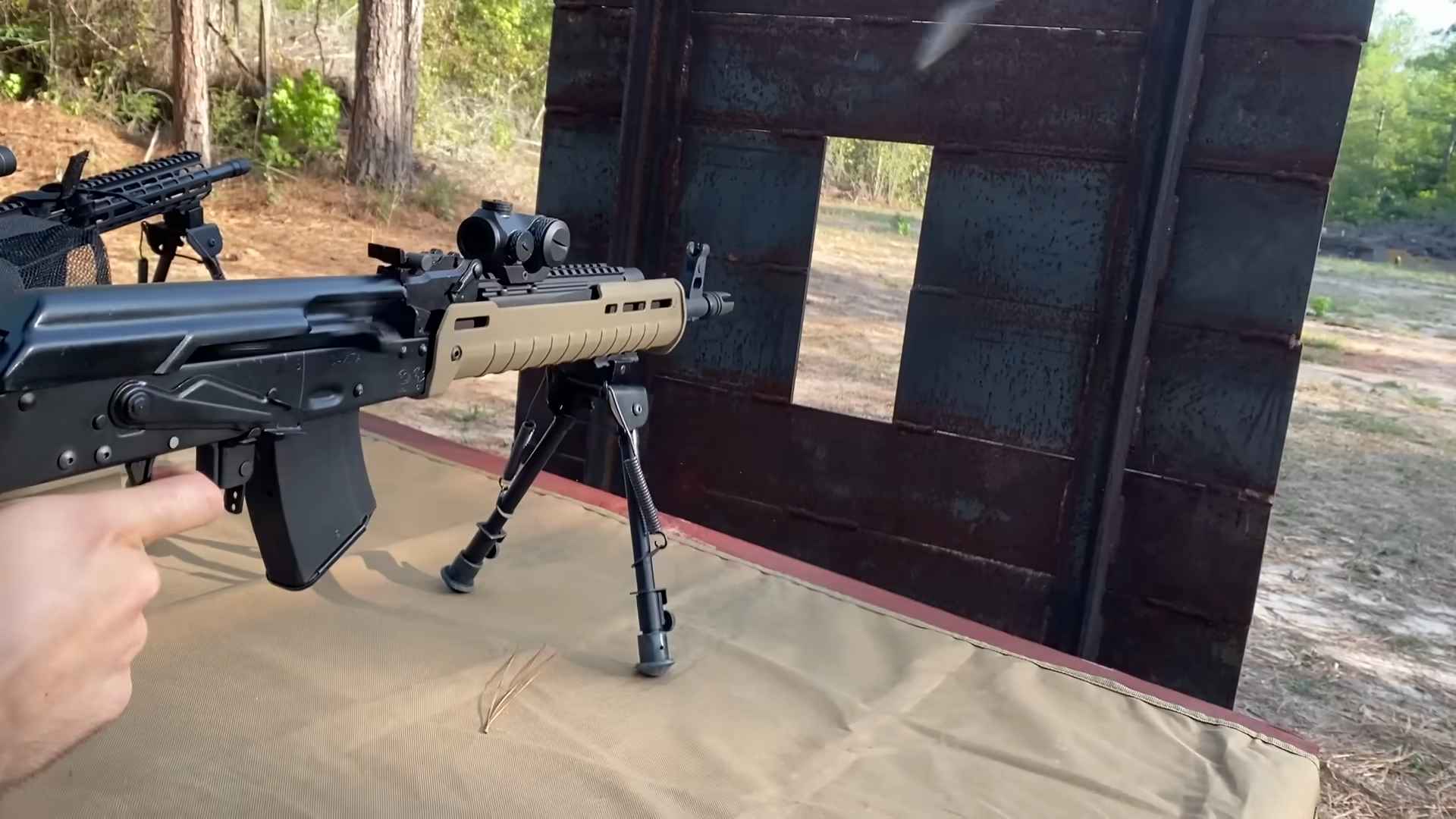 Shooter aiming a rifle from a bench rest setup, firing through a steel shooting barrier with a visible muzzle flash