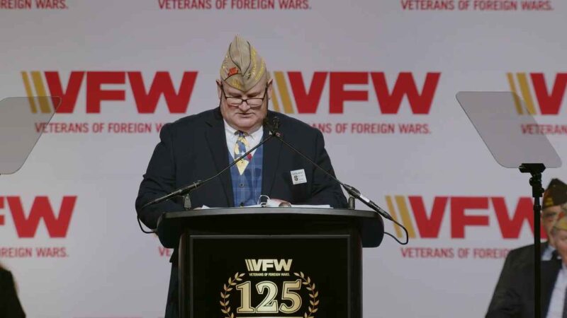 A speaker at a podium during the 125th VFW National Convention in Phoenix, wearing a VFW cap and formal attire, with the Veterans of Foreign Wars logo in the background