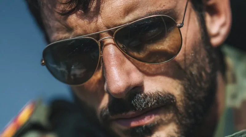 Close-up of a man wearing aviator sunglasses, reflecting a confident look with a military-style jacket visible
