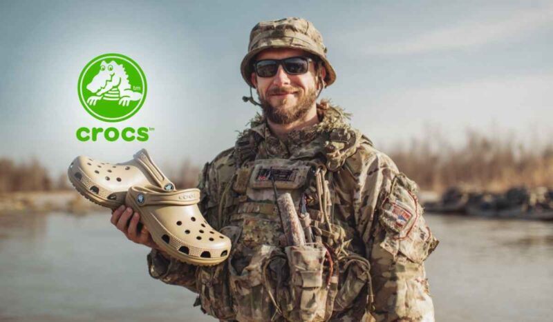 A smiling soldier in camouflage uniform holding a pair of tan Crocs, with the Crocs logo in the background