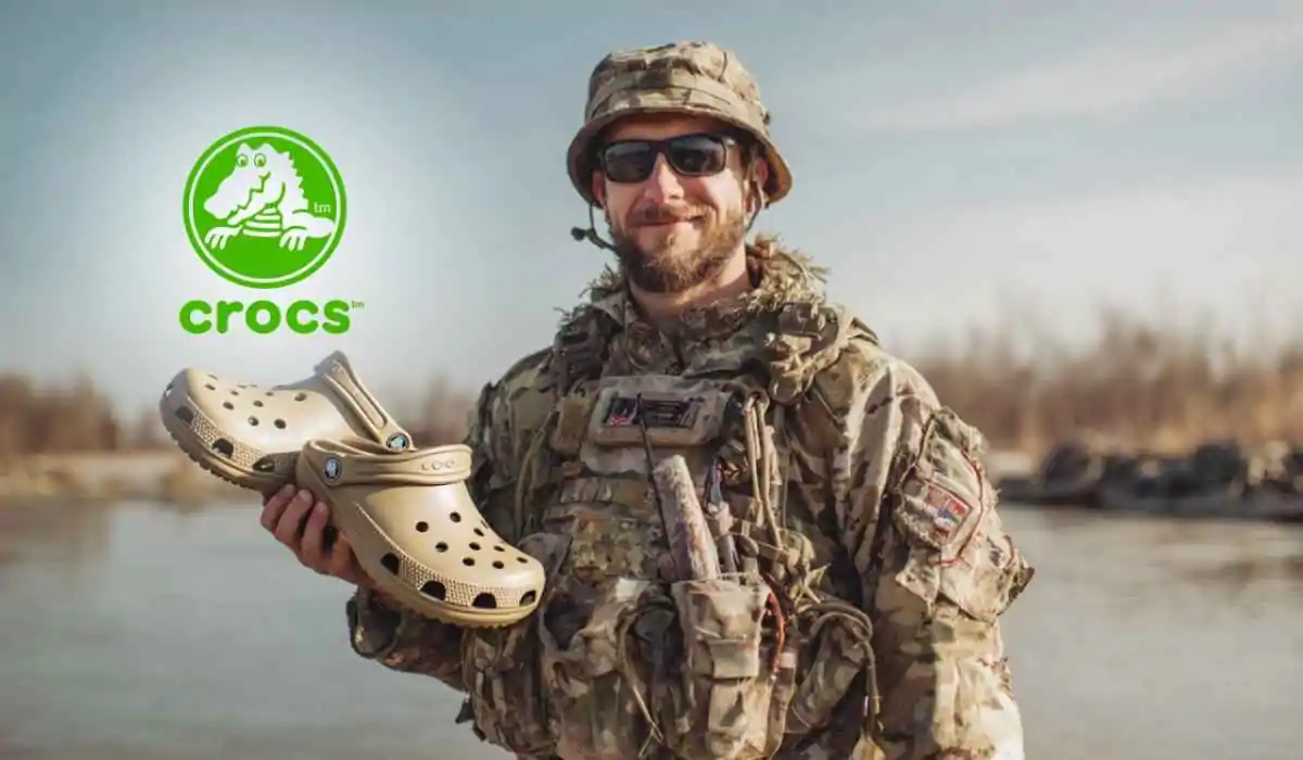 A smiling soldier in camouflage uniform holding a pair of tan Crocs, with the Crocs logo in the background