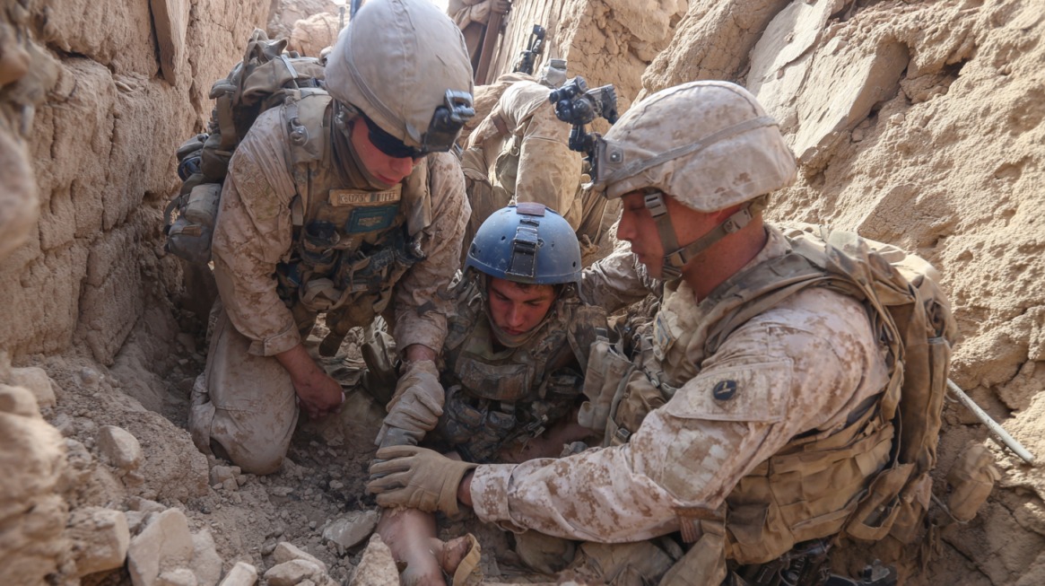 Two soldiers in tan uniforms assist an injured fellow soldier sitting in a dirt trench