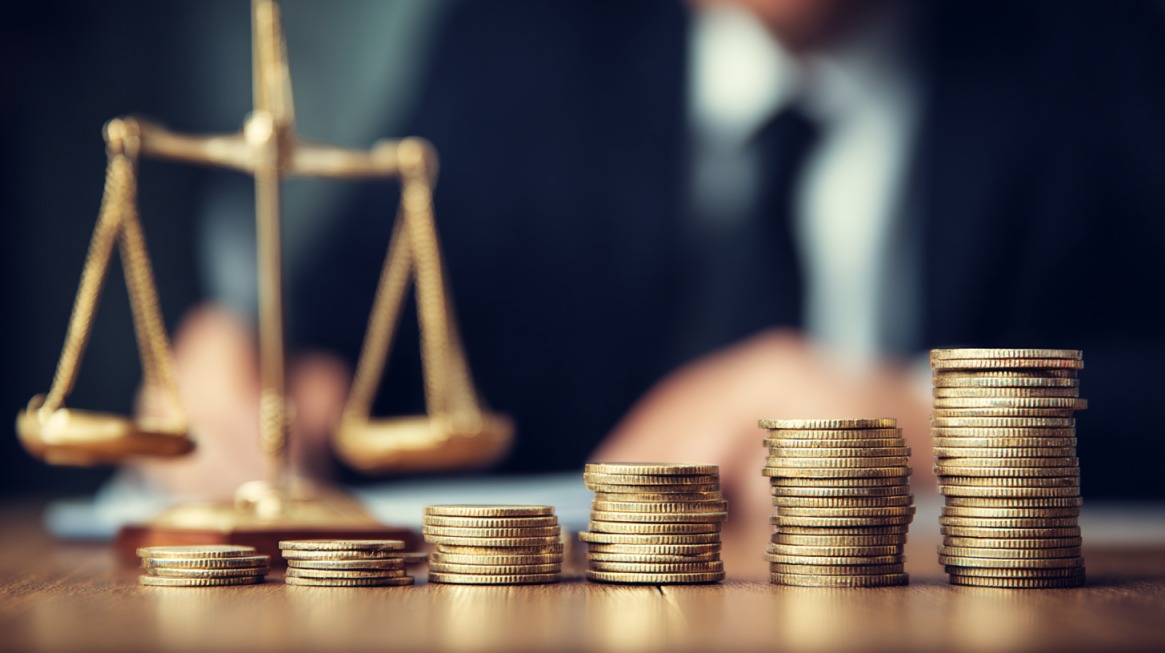 Close-up of stacked gold coins in front of golden scales of justice, with a blurred person in a suit in the background