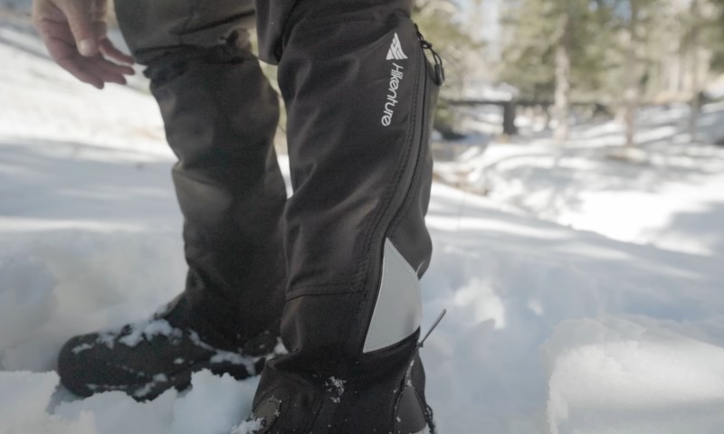 Close-up of a person wearing black water-resistant gaiters branded "Venture", standing in deep snow