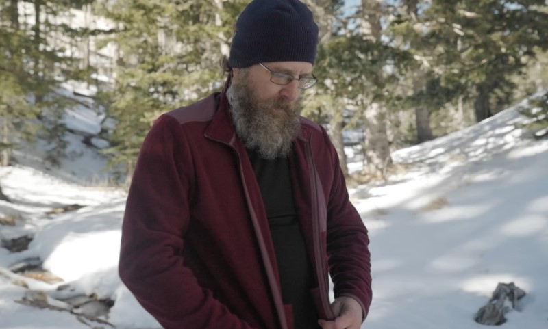 A bearded man in a red jacket and navy beanie stands in a snowy forest